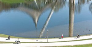 Ein See in dem sich Gebäudeteile spiegeln, am Rand Fussgänger und Radfahrer