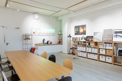 Atelier-dieSmartphotoschule Eine Frau mit braunem, welligem Haar und roter Brille hält ein Smartphone, das an einem Gimbal befestigt ist, und nimmt ein Video auf. Im Hintergrund sind verschiedene Bilder an einer weißen Wand angebracht. Der Fokus liegt auf der Videoproduktion, während sie möglicherweise eine Anleitung oder Präsentation aufzeichnet. Die Szene deutet auf eine kreative und professionelle Umgebung hin, in der Video-Content erstellt wird.