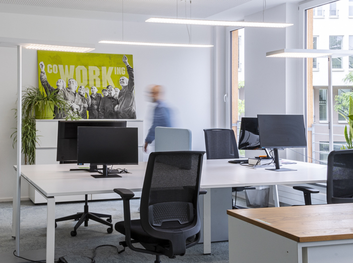 Workshop-SmartphoneFotografie-für_Business Ein moderner, heller Büroraum mit mehreren leeren Schreibtischen und Bürostühlen. Auf den Schreibtischen stehen Computerbildschirme, und im Hintergrund hängt ein großes Poster mit der Aufschrift 'CO-WORKING' und einem Bild von Personen. Eine unscharfe Gestalt im blauen Hemd geht durch den Raum, was Bewegung andeutet. Große Fenster lassen viel Tageslicht herein.