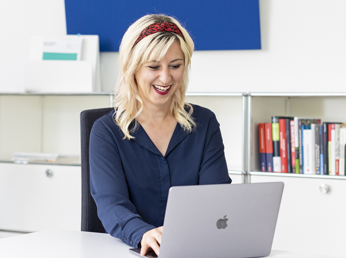 Workshop Smartphone Fotografie für Business Eine blonde Frau mit einem rot-schwarzen Haarband sitzt lächelnd an einem Tisch und arbeitet an einem Laptop. Sie trägt eine dunkelblaue Bluse. Im Hintergrund befinden sich ein Regal mit Büchern und eine weiße Kommode, darüber hängt ein blaues Wandpaneel.