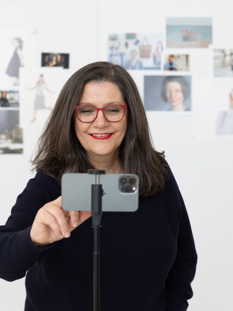Training_Smartphone_Fotografie_Business Frau mit langen dunklen Haaren und Brille fotografiert sich mit dem Smartphone auf einem Stativ. Im Hintergrund eine Wand mit ausgedruckten Porträts und Fotobeispielen. Symbolbild für den Workshop „Professionelle Smartphone-Fotografie.