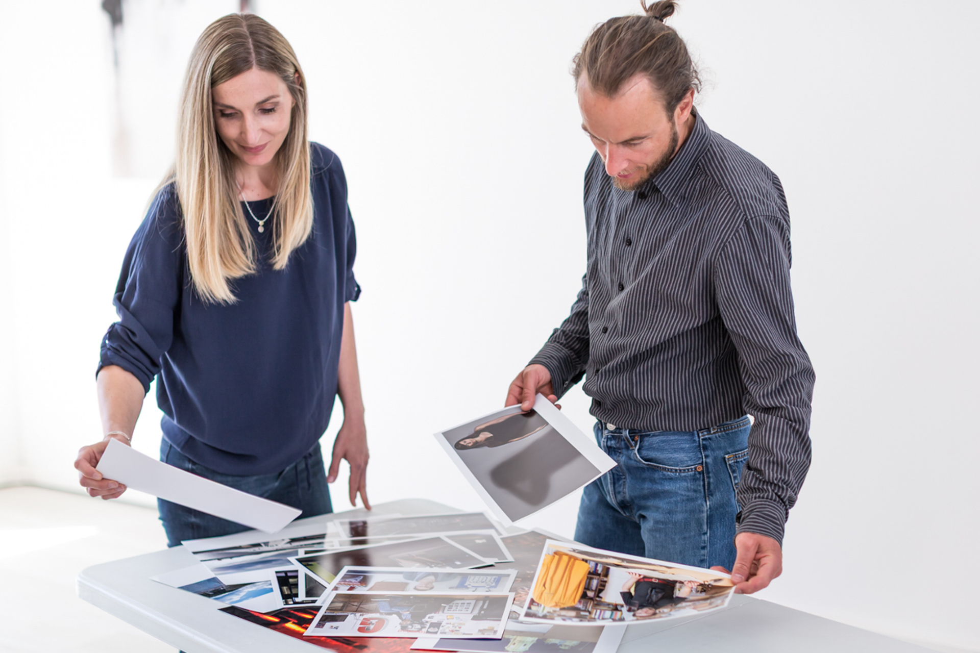 Eine Frau und ein Mann stehen an einem weißen Tisch und betrachten gemeinsam eine Auswahl an ausgedruckten Fotos. Beide halten jeweils ein Bild in der Hand und wirken konzentriert. Die helle Umgebung und die vielfältigen Motive auf dem Tisch deuten auf einen kreativen Prozess zur Entwicklung einer Bildsprache oder zur Bildauswahl hin.