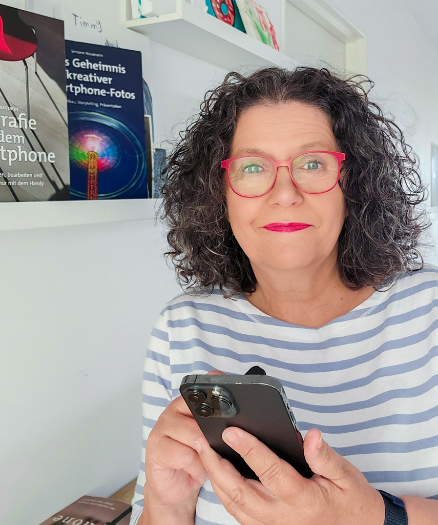 Eine Frau mit lockigem, dunklem Haar und roter Brille steht in einem hellen Raum und hält ein Smartphone in den Händen. Sie trägt ein weiß-blau gestreiftes Shirt und schaut direkt in die Kamera. Im Hintergrund sind auf einem weißen Regal mehrere Bücher zu sehen, darunter eines mit dem Titel „Das Geheimnis kreativer Smartphone-Fotos.