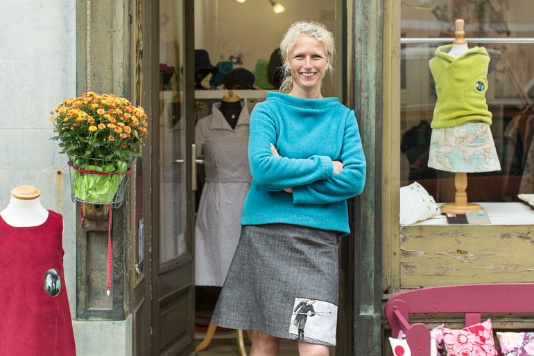 Eine Frau mit lockigem, grauem Haar steht lächelnd vor ihrem kleinen Laden. Sie trägt einen türkisfarbenen Pullover und einen grauen Rock. Neben ihr sind ein Blumenarrangement und Schaufensterpuppen mit handgefertigter Kleidung zu sehen.