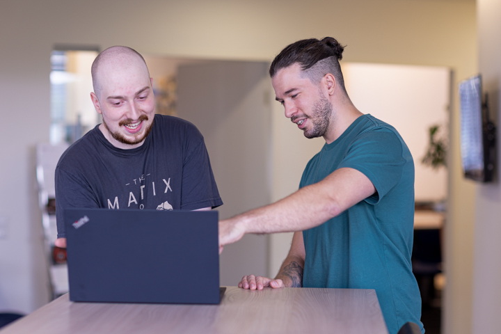 aptop im Vordergrund, zwei Männer diskutieren dahinter am Bildschirm – Beispiel für Businessfotografie und visuelle Kommunikation im digitalen Arbeitsalltag