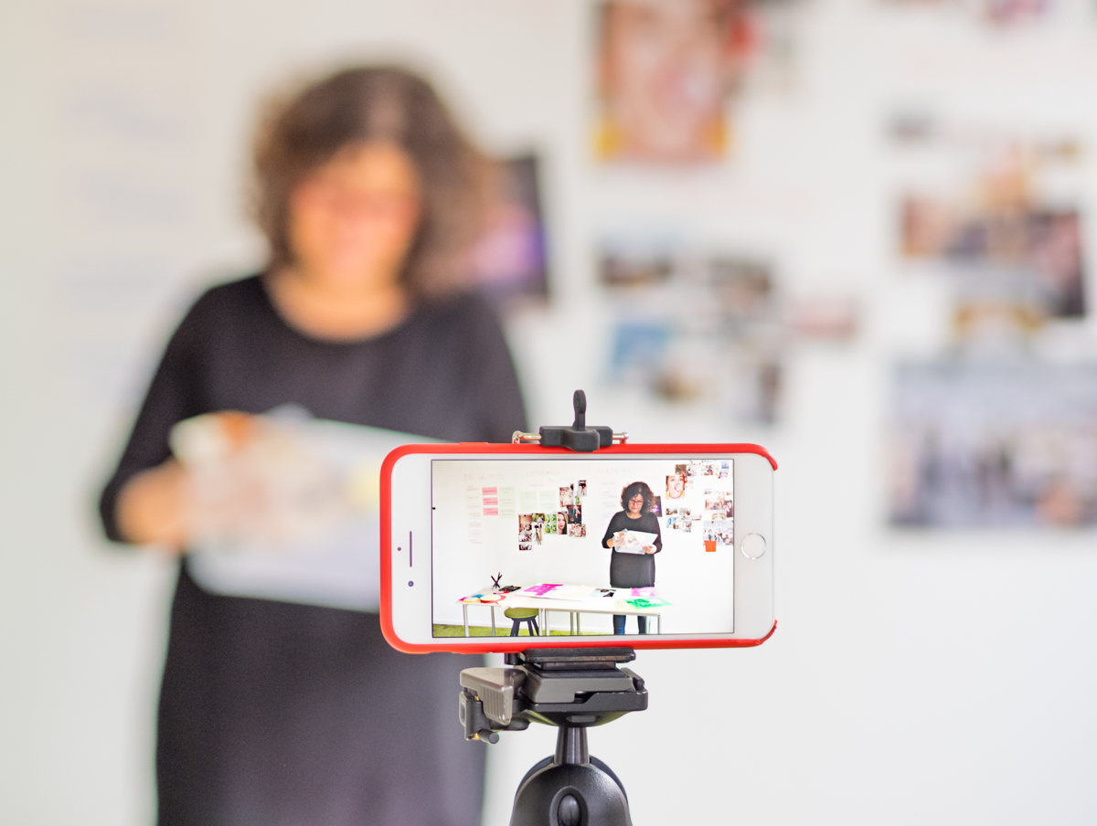 Ein Smartphone filmt eine kreative Bildmacherin bei der Arbeit vor einer Wand mit Bildbeispielen und visuellen Ideen. Die Szene zeigt, wie Bildsprache, Smartphone-Video und kreatives Erzählen zusammenkommen, um Inhalte sichtbar, lebendig und strategisch wirksam zu machen.