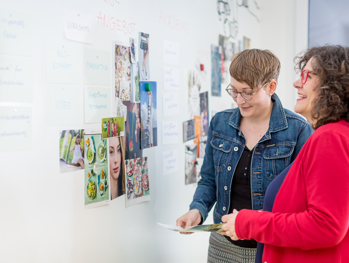 Zwei Frauen stehen vor einer Wand mit Moodboards und Notizen und betrachten gemeinsam Bildmaterial. Die Szene zeigt einen kreativen Austausch über Bildsprache, visuelle Wirkung und die gezielte Auswahl von Motiven für Kommunikation und Marketing.