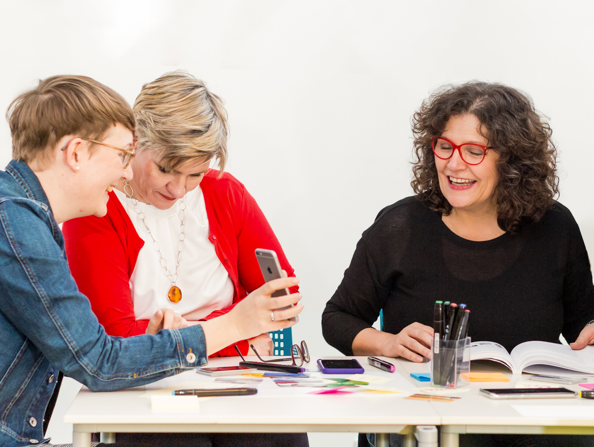 Drei Frauen sitzen gemeinsam an einem Tisch und arbeiten praktisch mit Smartphone, Notizen und Bildmaterial. Die Szene zeigt einen lebendigen Workshop zu Bildsprache und visueller Strategie, bei dem Ideen direkt ausprobiert, besprochen und in konkrete Kommunikation übersetzt werden.